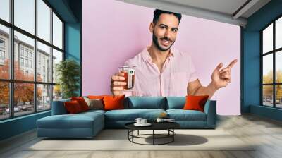 Young hispanic man holding cup of tea smiling happy pointing with hand and finger to the side Wall mural