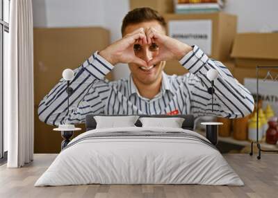 Young hispanic man making heart shape with hands in a warehouse setting, wearing striped shirt. Wall mural