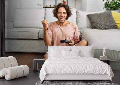 Young hispanic man playing video game holding controller at home screaming proud, celebrating victory and success very excited with raised arms Wall mural