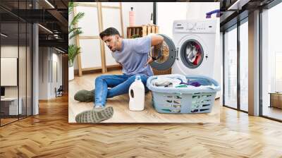 Young hispanic man putting dirty laundry into washing machine suffering of backache, touching back with hand, muscular pain Wall mural