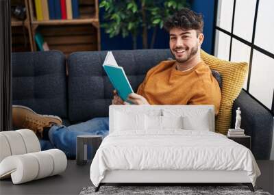 Young hispanic man reading book sitting on sofa at home Wall mural