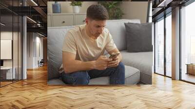 Young hispanic man sitting on a couch in his living room, focused on his phone in a modern, cozy apartment. Wall mural