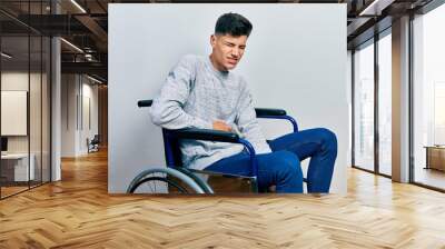 Young hispanic man sitting on wheelchair with hand on stomach because indigestion, painful illness feeling unwell. ache concept. Wall mural