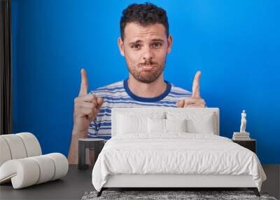 Young hispanic man standing over blue background pointing up looking sad and upset, indicating direction with fingers, unhappy and depressed. Wall mural