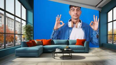 Young hispanic man standing over blue background relaxed and smiling with eyes closed doing meditation gesture with fingers. yoga concept. Wall mural