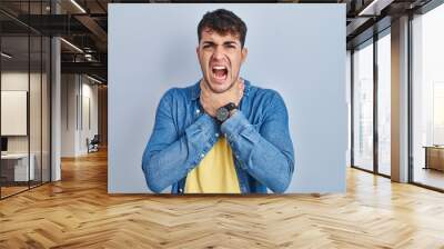 Young hispanic man standing over blue background shouting suffocate because painful strangle. health problem. asphyxiate and suicide concept. Wall mural