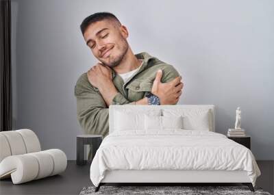 Young hispanic man standing over isolated background hugging oneself happy and positive, smiling confident. self love and self care Wall mural