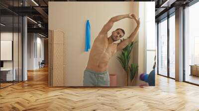 Young hispanic man stretching in a gym room with indoor plants and fitness equipment including exercise balls and a wooden partition in the background Wall mural