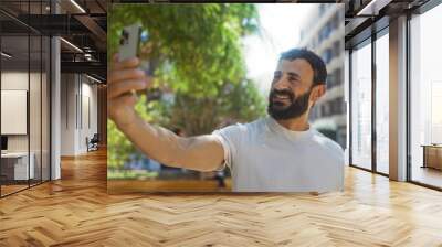 Young hispanic man takes a selfie in an urban park with a modern cityscape in the background Wall mural