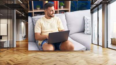 Young hispanic man using laptop sitting on sofa at home Wall mural