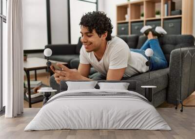 Young hispanic man using smartphone lying on the sofa at home. Wall mural