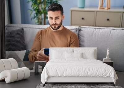 Young hispanic man using smartphone sitting on sofa at home Wall mural