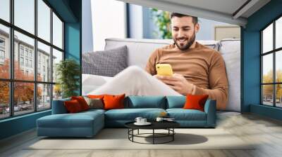 Young hispanic man using smartphone sitting on sofa at home Wall mural