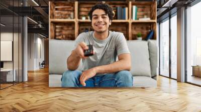 Young hispanic man watching tv sitting on the sofa at home. Wall mural