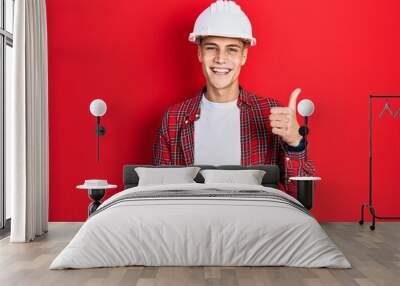 Young hispanic man wearing architect hardhat smiling happy and positive, thumb up doing excellent and approval sign Wall mural