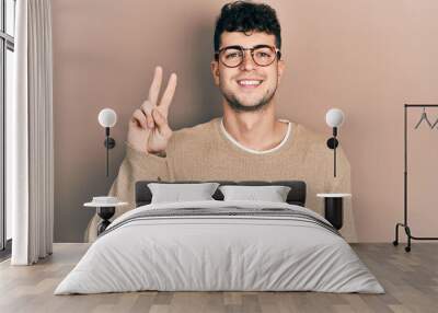 Young hispanic man wearing casual clothes and glasses smiling looking to the camera showing fingers doing victory sign. number two. Wall mural