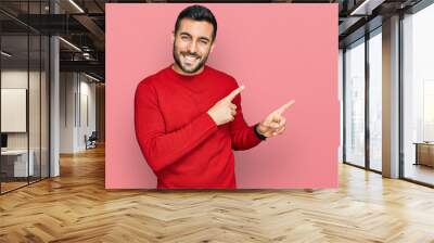 Young hispanic man wearing casual clothes smiling and looking at the camera pointing with two hands and fingers to the side. Wall mural