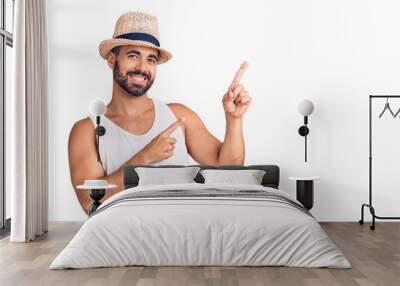 Young hispanic man wearing casual summer hat smiling and looking at the camera pointing with two hands and fingers to the side. Wall mural