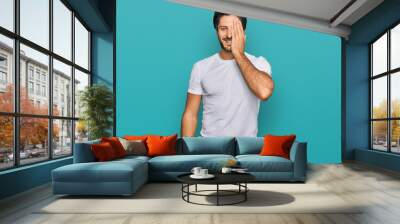 Young hispanic man wearing casual white t shirt covering one eye with hand, confident smile on face and surprise emotion. Wall mural