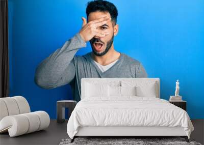 Young hispanic man wearing casual white t shirt peeking in shock covering face and eyes with hand, looking through fingers afraid Wall mural