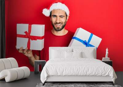 Young hispanic man wearing christmas hat and holding gifts winking looking at the camera with sexy expression, cheerful and happy face. Wall mural