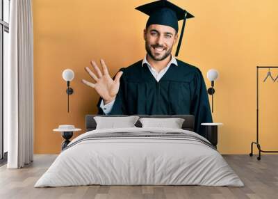 Young hispanic man wearing graduation cap and ceremony robe showing and pointing up with fingers number five while smiling confident and happy. Wall mural