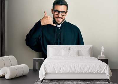 Young hispanic man wearing priest uniform standing over white background smiling doing phone gesture with hand and fingers like talking on the telephone. communicating concepts. Wall mural