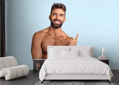 Young hispanic man wearing swimwear shirtless with a big smile on face, pointing with hand and finger to the side looking at the camera. Wall mural