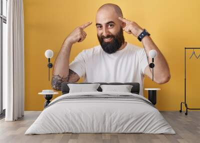 Young hispanic man with beard and tattoos standing over yellow background smiling pointing to head with both hands finger, great idea or thought, good memory Wall mural