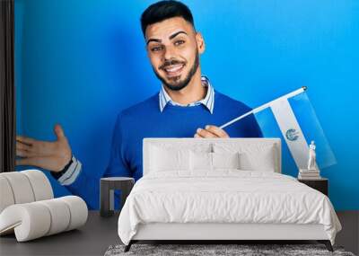 Young hispanic man with beard holding el salvador flag celebrating achievement with happy smile and winner expression with raised hand Wall mural