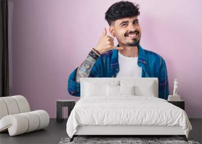 Young hispanic man with beard standing over pink background smiling doing phone gesture with hand and fingers like talking on the telephone. communicating concepts. Wall mural