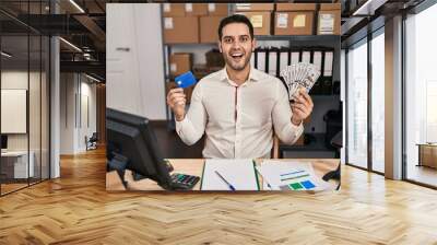 Young hispanic man with beard working at small business ecommerce holding banknotes and credit card celebrating crazy and amazed for success with open eyes screaming excited. Wall mural