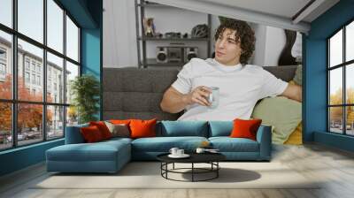 Young hispanic man with curly hair in a casual white t-shirt holding a mug, leisurely reclining on a gray sofa with colorful pillows in a stylish home Wall mural