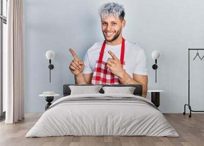 Young hispanic man with modern dyed hair wearing apron smiling and looking at the camera pointing with two hands and fingers to the side. Wall mural