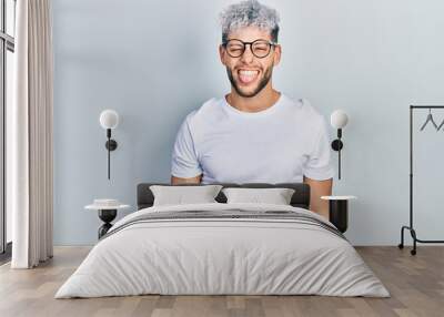 Young hispanic man with modern dyed hair wearing white t shirt and glasses sticking tongue out happy with funny expression. emotion concept. Wall mural