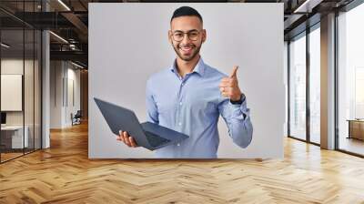 Young hispanic man working using computer laptop doing happy thumbs up gesture with hand. approving expression looking at the camera showing success. Wall mural