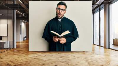 Young hispanic priest man holding bible in shock face, looking skeptical and sarcastic, surprised with open mouth Wall mural