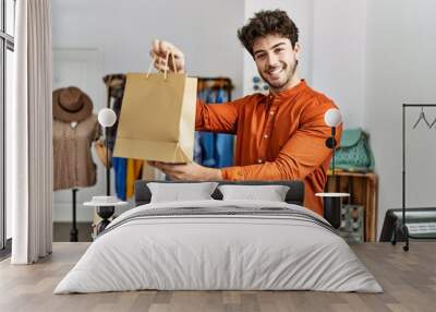 Young hispanic shopkeeper man smiling happy holding shopping bag at clothing store. Wall mural