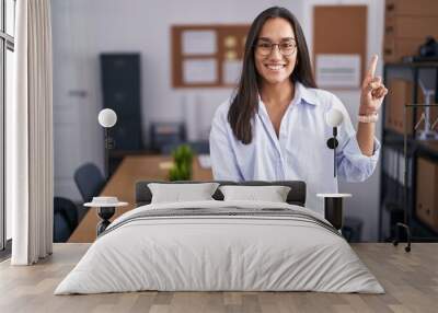 Young hispanic woman at the office showing and pointing up with finger number one while smiling confident and happy. Wall mural