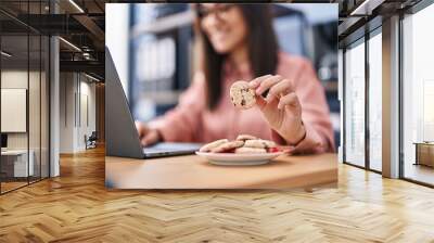 Young hispanic woman business worker using laptop eating cookies at office Wall mural