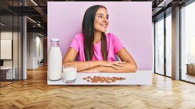 Young hispanic woman drinking healthy almond milk sitting on the table looking to side, relax profile pose with natural face and confident smile. Wall mural