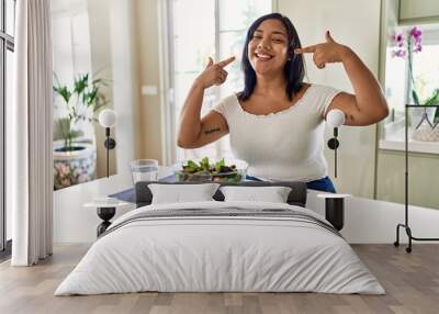 Young hispanic woman eating healthy salad at home smiling cheerful showing and pointing with fingers teeth and mouth. dental health concept. Wall mural