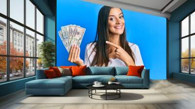 Young hispanic woman holding dollars smiling happy pointing with hand and finger Wall mural