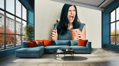 Young hispanic woman holding ice cream angry and mad screaming frustrated and furious, shouting with anger. rage and aggressive concept. Wall mural