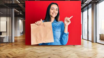 Young hispanic woman holding take away paper bag smiling happy pointing with hand and finger to the side Wall mural