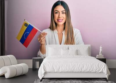 Young hispanic woman holding venezuelan flag sitting on the table looking positive and happy standing and smiling with a confident smile showing teeth Wall mural