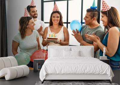 Young hispanic woman smiling happy and holding birthday cake standing with a group of friends at home. Wall mural