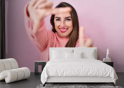 Young hispanic woman standing over pink background smiling making frame with hands and fingers with happy face. creativity and photography concept. Wall mural