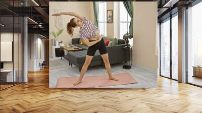 Young hispanic woman stretching on a yoga mat in a bright living room, depicting an indoor fitness routine. Wall mural