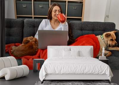 Young hispanic woman using laptop and drinking coffee sitting on sofa with dogs at home Wall mural